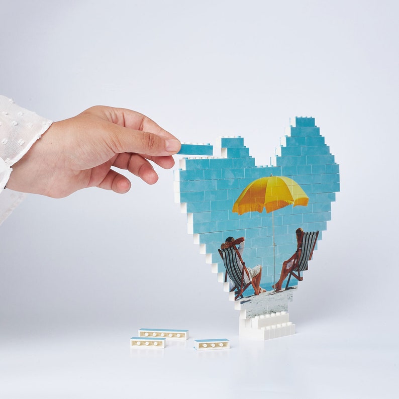 May include: A blue and white heart-shaped mosaic with a picture of a beach scene. The scene includes two people sitting in chairs under a yellow umbrella. The mosaic is made of small plastic bricks.