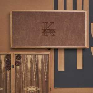 May include: A wooden backgammon game set with a brown leather-like lid. The lid has a monogrammed "K" with the name "KINGSTON" underneath. The game board is made of wood with a light brown and dark brown wood inlay design.