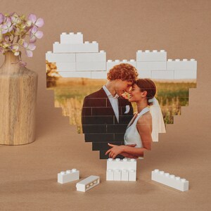 May include: A white LEGO heart frame with a photo of a couple in love. The couple is standing in a field and looking at each other. The photo is surrounded by white LEGO bricks.