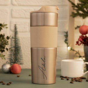 May include: A gold-coloured stainless steel travel mug with a beige lid and a black inscription that reads "Adela".