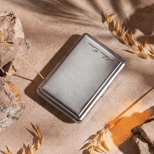 May include: A silver cigarette case with the name "Adrian Evans" in cursive script. The case has a brushed metal finish and a rectangular shape with rounded corners. The background is a neutral beige with decorative elements.