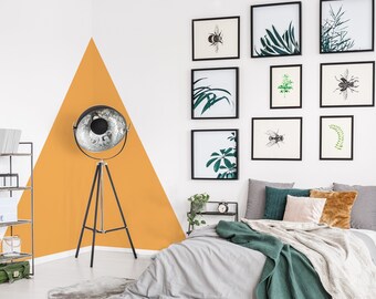 Puede incluir: Un dormitorio con una pared blanca y amarilla, una cama con una manta gris y verde, y una lámpara de trípode negra con un reflector plateado. La pared está decorada con estampados enmarcados de plantas e insectos.