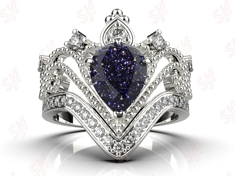 May include: A silver ring with a large blue pear-shaped gemstone surrounded by smaller clear gemstones. The ring is designed in a crown shape with intricate details.