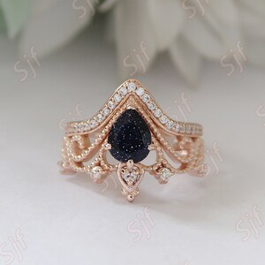 May include: A rose gold ring with a pear-shaped blue gemstone surrounded by smaller clear gemstones. The ring has a delicate, intricate design with a crown-like shape.