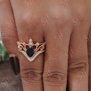 May include: A rose gold ring with a black heart-shaped stone surrounded by smaller clear stones. The ring is designed in a crown shape with intricate details.