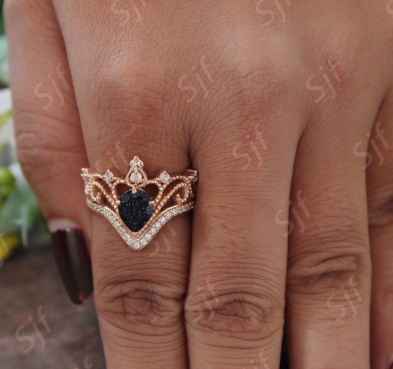 May include: A rose gold ring with a black heart-shaped stone surrounded by smaller clear stones. The ring is designed in a crown shape with intricate details.