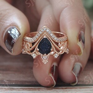 May include: A rose gold ring with a black pear-shaped stone surrounded by clear stones. The ring has a delicate, intricate design with a crown-like top.