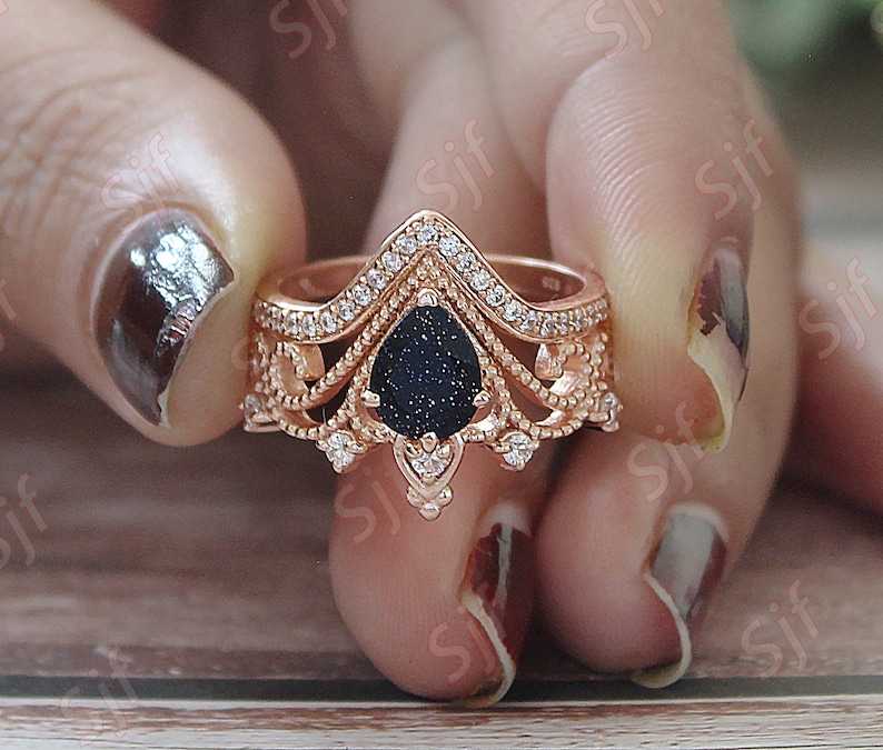 May include: A rose gold ring with a black pear-shaped stone surrounded by clear stones. The ring has a delicate, intricate design with a crown-like top.