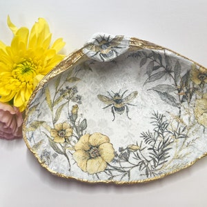 May include: A white ceramic shell dish with a gold rim and a floral design featuring yellow flowers and bees. The dish is decorated with a delicate, hand-painted design.