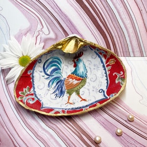 May include: A red and white ceramic dish shaped like a seashell with a gold rim. The dish features a colorful rooster design and is decorated with floral patterns. Three white pearls are placed on a pink and white marbled surface.