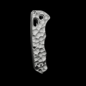 May include: A silver knife handle with a textured, honeycomb pattern. The handle features several holes and a curved design for grip. The background is black, accentuating the metallic finish and intricate details of the knife handle.