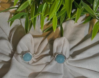 May include: Two silver rings with light blue gemstone accents. The rings are on a white fabric background.