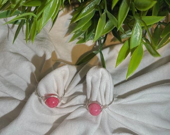 Könnte beinhalten: Zwei silberne Ringe mit rosafarbenen Edelsteinperlen. Die Ringe liegen auf einem weißen Stoffhintergrund.