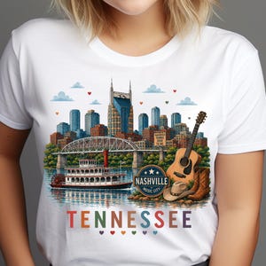 May include: A white t-shirt featuring a colorful graphic print of the Nashville skyline, a riverboat, an acoustic guitar, and cowboy boots. The design includes the text Nashville Music City and Tennessee with small heart icons below the word.