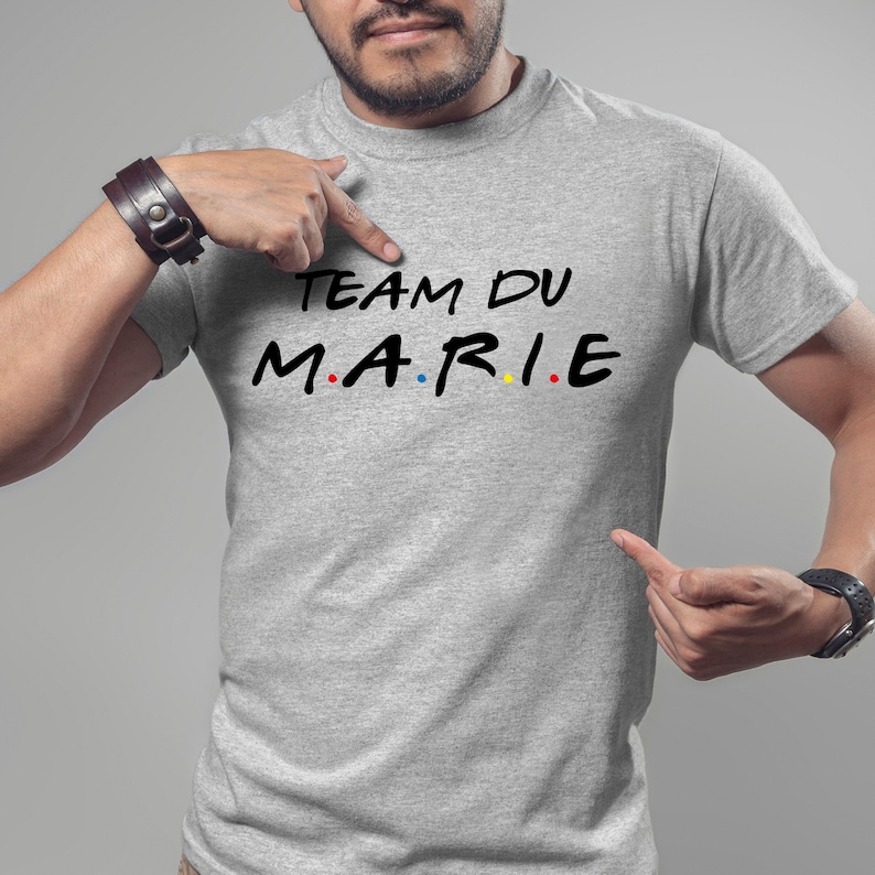 K&ouml;nnte beinhalten: Ein hellgraues T-Shirt mit dem Text "TEAM DU MARIE" in Schwarz, wobei das Wort "MARIE" im Stil der Fernsehserie "Friends" gestaltet ist. Die Buchstaben sind in Schwarz, Rot, Blau, Gelb und Rot.