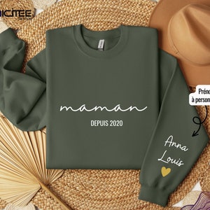 Könnte beinhalten: Olivgrünes Sweatshirt mit dem Wort "maman" und "DEPUIS 2020" in weißer Schrift. Der Ärmel zeigt die Namen "Anna Louis" und ein goldenes Herz. Das Sweatshirt ist von der Marke Gildan.