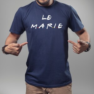 K&ouml;nnte beinhalten: Marineblaues T-Shirt mit dem wei&szlig;en Schriftzug "LE MARIE", wobei die Buchstaben "MARIE" im Stil der Fernsehserie "Friends" gehalten sind. Die Buchstaben haben rote, blaue und gelbe Punkte. Eine Person zeigt auf das T-Shirt.