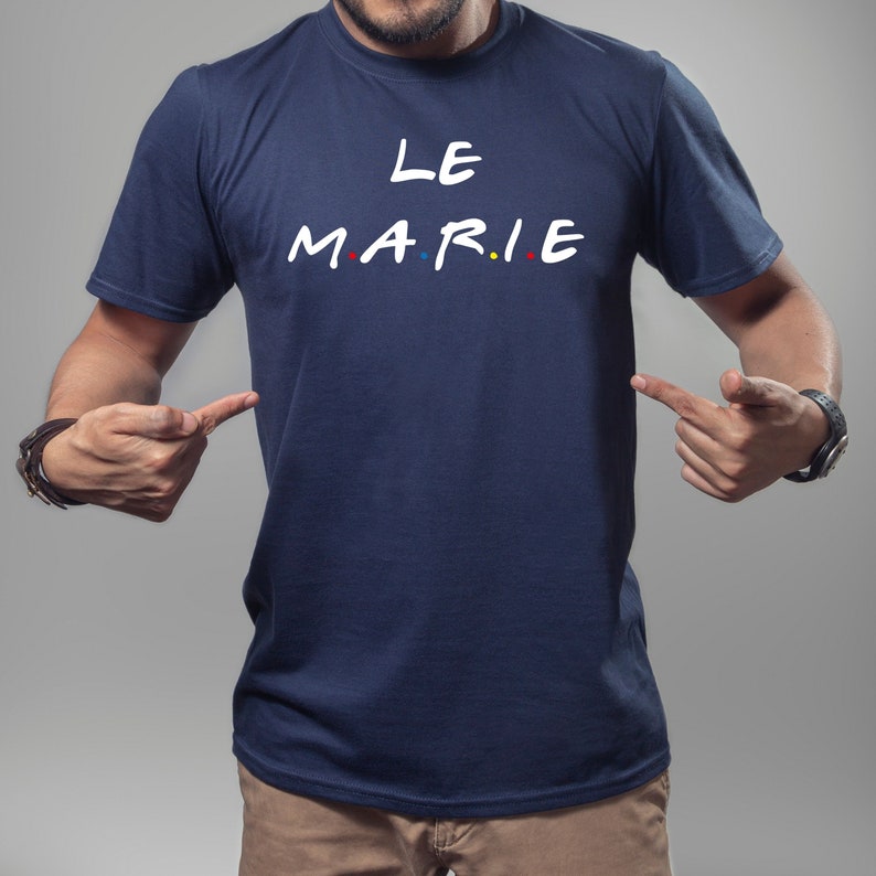K&ouml;nnte beinhalten: Marineblaues T-Shirt mit dem wei&szlig;en Schriftzug "LE MARIE", wobei die Buchstaben "MARIE" im Stil der Fernsehserie "Friends" gehalten sind. Die Buchstaben haben rote, blaue und gelbe Punkte. Eine Person zeigt auf das T-Shirt.