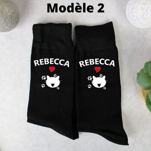 Op de afbeelding: Een paar zwarte sokken met een wit kattenontwerp en de naam "Rebecca" gedrukt op elke sok. Het kattenontwerp bevat een rood hart.