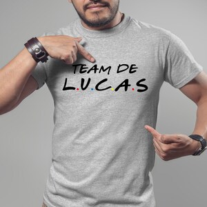 K&ouml;nnte beinhalten: Hellgraues T-Shirt mit dem schwarzen Text "TEAM DE LUCAS", wobei die Buchstaben "L.U.C.A.S" im Stil der Fernsehserie "Friends" gehalten sind. Eine Person zeigt auf das T-Shirt.