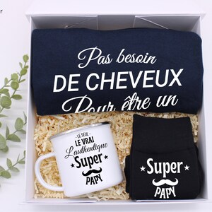 May include: A gift box set for a new dad. The set includes a navy blue t-shirt with the text "Pas besoin de cheveux pour &ecirc;tre un", a black mug with the text "Le seul, le vrai, l'authentique *Super* Papy", and black socks with the text "*Super* Papy" and a mustache graphic.