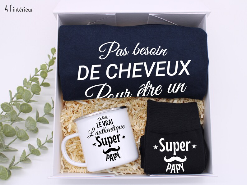 May include: A gift box set for a new dad. The set includes a navy blue t-shirt with the text "Pas besoin de cheveux pour &ecirc;tre un", a black mug with the text "Le seul, le vrai, l'authentique *Super* Papy", and black socks with the text "*Super* Papy" and a mustache graphic.
