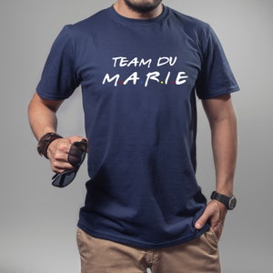 K&ouml;nnte beinhalten: Marineblaues T-Shirt mit dem wei&szlig;en Schriftzug "TEAM DU MARIE", wobei das Wort "MARIE" im Stil der Fernsehserie "Friends" gestaltet ist. Das T-Shirt hat einen Rundhalsausschnitt und kurze &Auml;rmel.