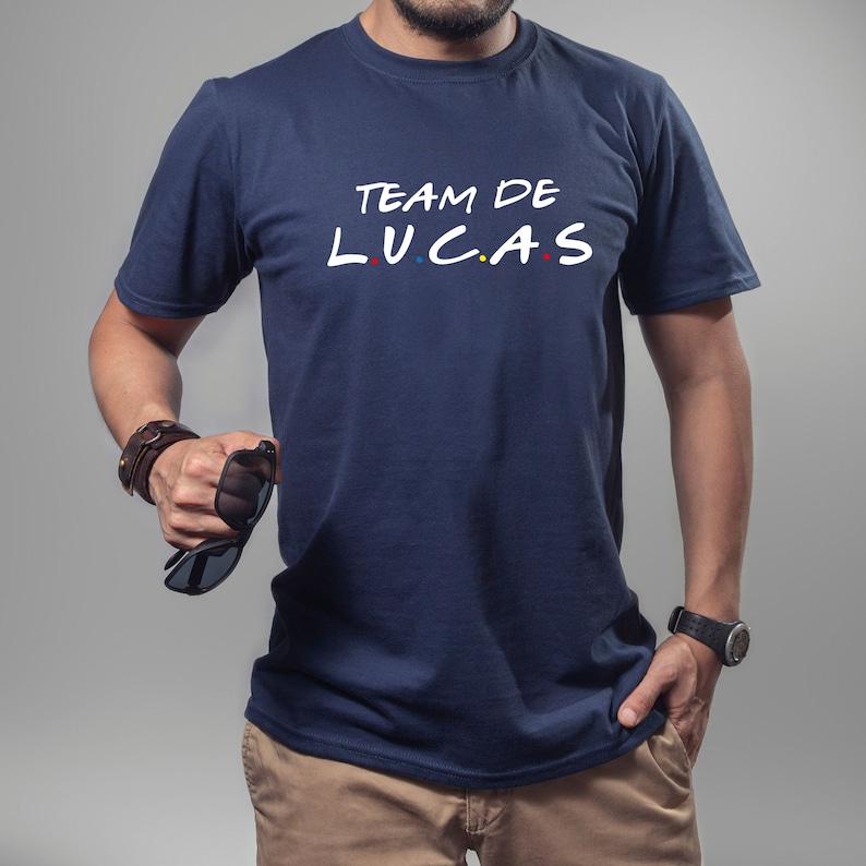 K&ouml;nnte beinhalten: Marineblaues T-Shirt mit dem wei&szlig;en Schriftzug "TEAM DE LUCAS", wobei das Wort "LUCAS" im Stil der Fernsehserie Friends geschrieben ist. Das Shirt hat einen Rundhalsausschnitt und kurze &Auml;rmel.