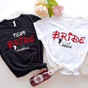 Op de afbeelding: Twee zwarte en witte T-shirts met een rood Disney-achtig lettertype dat "Team Bride" en "Bride" zegt met een Tinkerbell-silhouet. Het zwarte shirt zegt "Team Bride Anabelle" en het witte shirt zegt "Bride Adele".