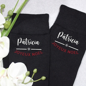 Op de afbeelding: Een paar zwarte sokken met witte tekst die "Patricia Joyeux Noel" en een sneeuwvlokje zegt.