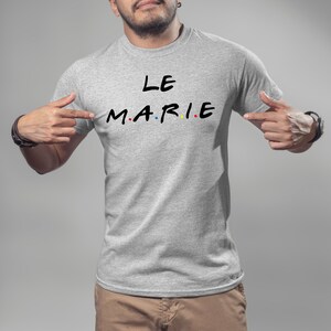 K&ouml;nnte beinhalten: Ein hellgraues T-Shirt mit dem Text "LE MARIE" in Schwarz, wobei die Buchstaben "M.A.R.I.E" im Stil der Fernsehserie "Friends" gehalten sind. Die Buchstaben sind rot, gelb und blau gef&auml;rbt. Das T-Shirt wird von einer Person gezeigt.