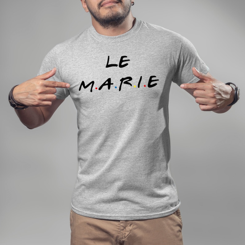 K&ouml;nnte beinhalten: Ein hellgraues T-Shirt mit dem Text "LE MARIE" in Schwarz, wobei die Buchstaben "M.A.R.I.E" im Stil der Fernsehserie "Friends" gehalten sind. Die Buchstaben sind rot, gelb und blau gef&auml;rbt. Das T-Shirt wird von einer Person gezeigt.