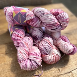 Pu&ograve; includere: Diversi gomitoli di filato tinto a mano nelle tonalit&agrave; del rosa, bianco e bordeaux. Il filato &egrave; esposto su una superficie di legno. Un'etichetta recita "Cable Tie Knits Hand Dyed Yarn". Il filato appare morbido ed &egrave; adatto per progetti di maglia o uncinetto.