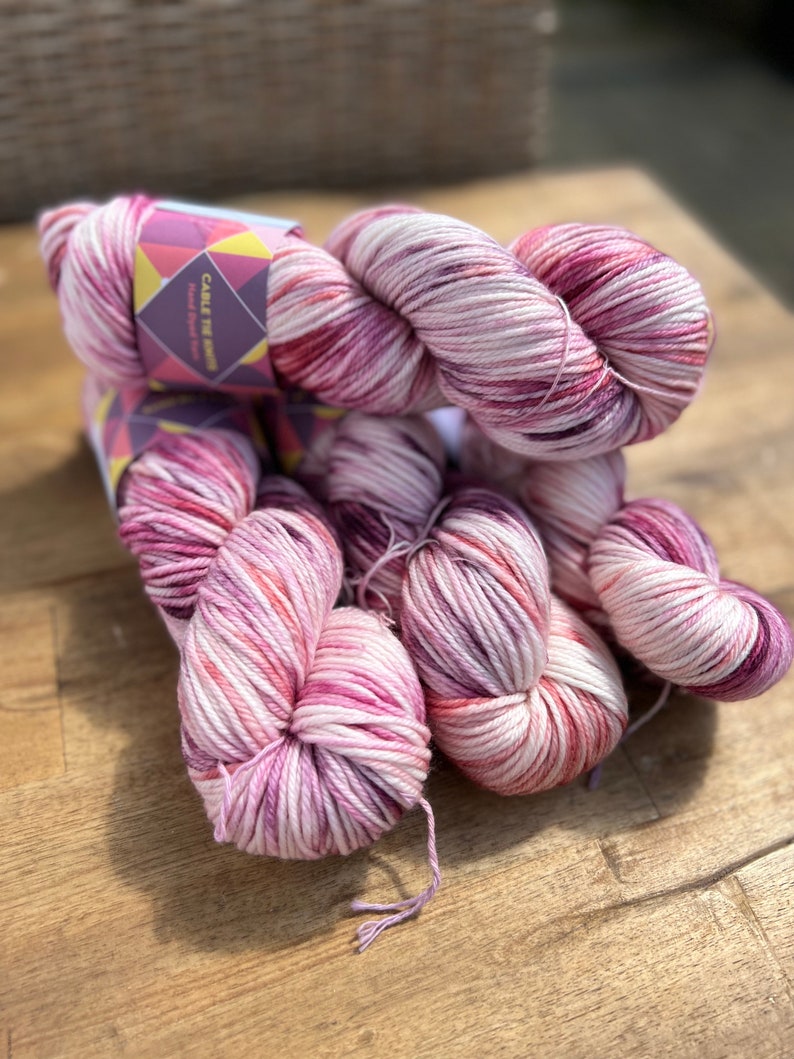 Pu&ograve; includere: Diversi gomitoli di filato tinto a mano nelle tonalit&agrave; del rosa, bianco e bordeaux. Il filato &egrave; esposto su una superficie di legno. Un'etichetta recita "Cable Tie Knits Hand Dyed Yarn". Il filato appare morbido ed &egrave; adatto per progetti di maglia o uncinetto.
