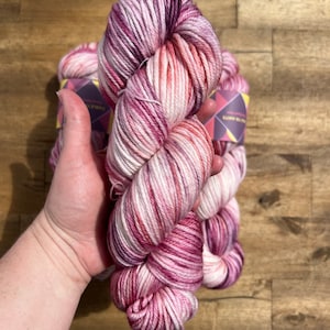Pu&ograve; includere: Un gomitolo di filato tinto a mano nei toni del rosa, bianco e magenta. Il filato &egrave; tenuto in una mano, con altri gomitoli visibili sullo sfondo. Il filato &egrave; etichettato "Cable Knits". Il filato &egrave; su una superficie di legno.