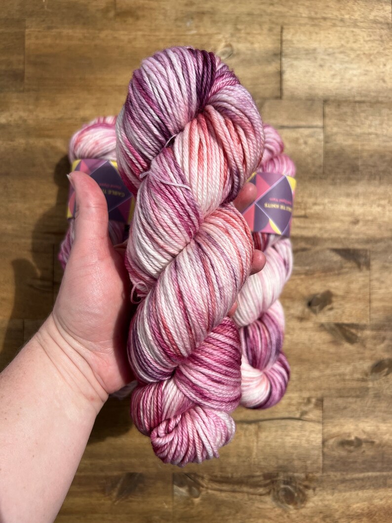 Pu&ograve; includere: Un gomitolo di filato tinto a mano nei toni del rosa, bianco e magenta. Il filato &egrave; tenuto in una mano, con altri gomitoli visibili sullo sfondo. Il filato &egrave; etichettato "Cable Knits". Il filato &egrave; su una superficie di legno.