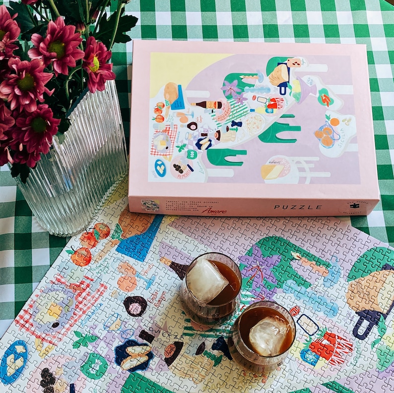 May include: A completed jigsaw puzzle with a colorful illustration of a picnic scene. Two glasses of iced coffee are on top of the puzzle. The puzzle box is in the background and says "Amore" and "Puzzle".