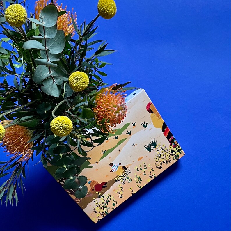 Puede incluir: Un ramo de flores amarillas y naranjas en una caja con una ilustraci&oacute;n colorida de personas y un paisaje des&eacute;rtico. La caja es de cart&oacute;n y tiene un fondo blanco.