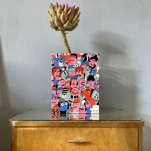 Op de afbeelding: Een kleurrijke illustratie van een menigte mensen met verschillende gezichtsuitdrukkingen en kapsels. De illustratie is gedrukt op een kaart met een witte achtergrond. De kaart ligt op een houten tafel.