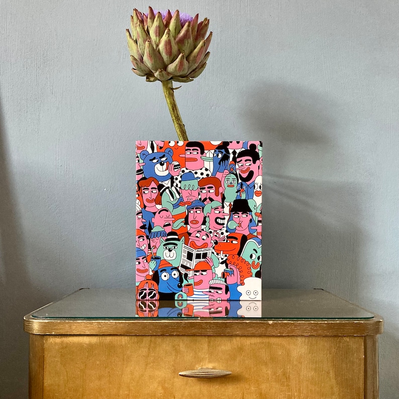 Op de afbeelding: Een kleurrijke illustratie van een menigte mensen met verschillende gezichtsuitdrukkingen en kapsels. De illustratie is gedrukt op een kaart met een witte achtergrond. De kaart ligt op een houten tafel.