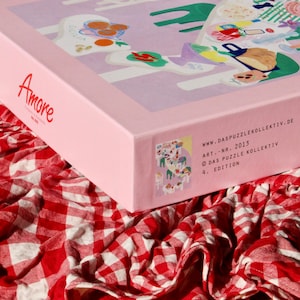 May include: A pink box with the word "Amore" on the front. The box is sitting on a red and white checkered tablecloth. The box has a colorful illustration of a picnic scene on the side. The text on the side reads "www.daspuzzlekollektiv.de ART.-NR. 2013 &copy; DAS PUZZLE KOLLEKTIV 4. EDITION"