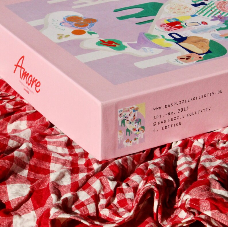 May include: A pink box with the word "Amore" on the front. The box is sitting on a red and white checkered tablecloth. The box has a colorful illustration of a picnic scene on the side. The text on the side reads "www.daspuzzlekollektiv.de ART.-NR. 2013 &copy; DAS PUZZLE KOLLEKTIV 4. EDITION"