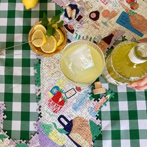 May include: A colorful jigsaw puzzle depicting an Italian scene with lemons, wine bottles, and a glass of lemonade. The puzzle is laid out on a green and white checkered tablecloth.