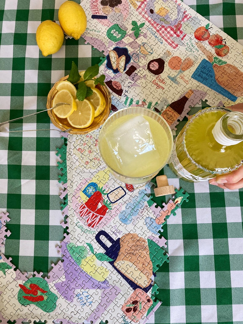 May include: A colorful jigsaw puzzle depicting an Italian scene with lemons, wine bottles, and a glass of lemonade. The puzzle is laid out on a green and white checkered tablecloth.