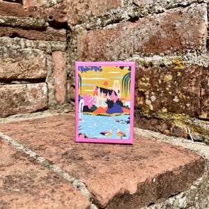 May include: A rectangular card with a bright pink border featuring a colorful illustration of a riverside scene. The artwork depicts a large building, a red sun, green trees, and two ducks swimming in blue water under a yellow sky.