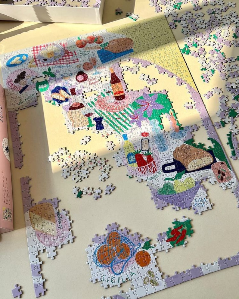 May include: A partially completed 1000-piece jigsaw puzzle with a colorful illustration of Italian food and produce. The puzzle pieces are on a light beige surface. The puzzle features a variety of Italian foods, including pasta, bread, tomatoes, oranges, and wine.