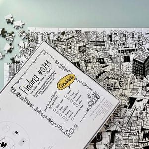 Puede incluir: Una caja blanca con texto negro que dice "Finding #07M" y una lista de verificaci&oacute;n de 11 cosas para encontrar en el rompecabezas. La caja est&aacute; encima de un rompecabezas en blanco y negro con una escena de ciudad.