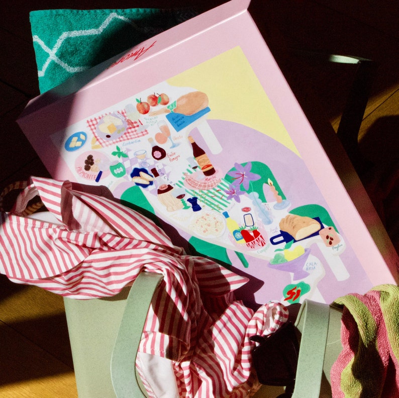 May include: A pink box with a colorful illustration of a map of Italy with various food items, including bread, cheese, and wine. The box is sitting on a green chair with a pink and white striped shirt draped over it.