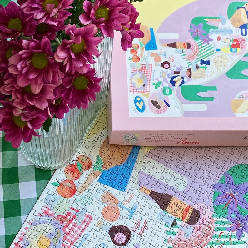 May include: A pink and green illustrated jigsaw puzzle with the word "Amore" on the box. The puzzle is laid out on a green and white checkered surface. A vase of pink flowers is in the background.