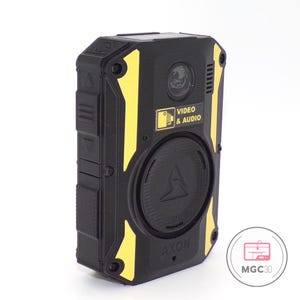 May include: A black body-worn camera with yellow accents, featuring a circular lens, a speaker grille, and a label reading VIDEO & AUDIO. The device has a textured circular button in the centre, side control buttons, and the word AXON printed at the bottom.